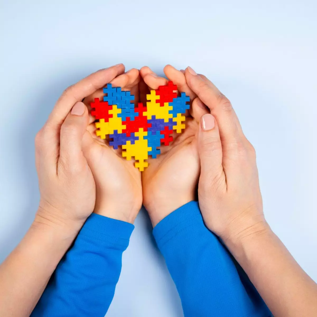 Imagem de mãos femininas e mãos de criança unidas, uma sobre a outra, segurando um coração formado por peças de quebra-cabeça coloridas, em alusão ao autismo.